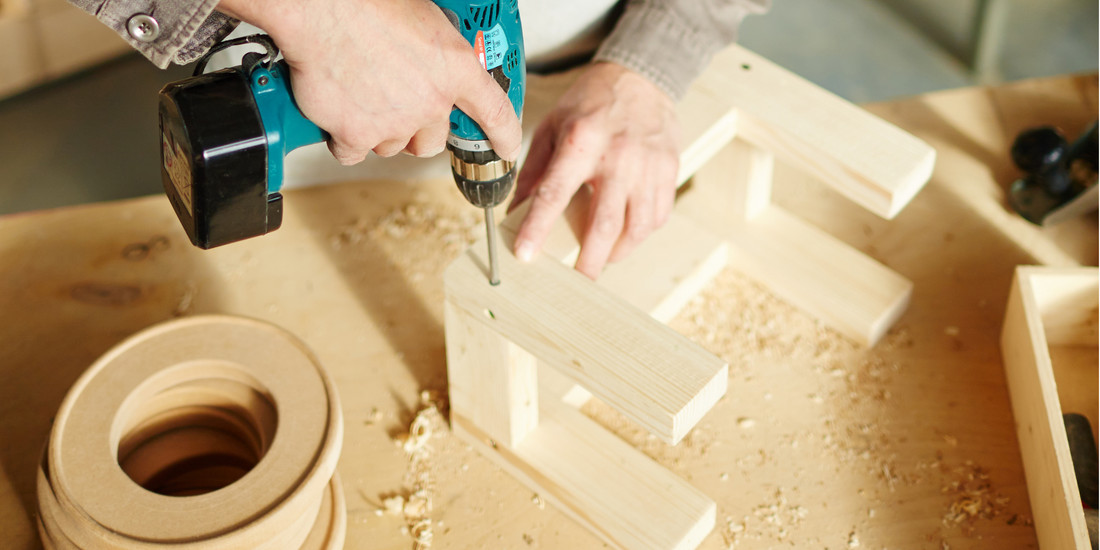 person using a teal electric drill to assemble wooden toys on a workbench with wood shavings scattered around.